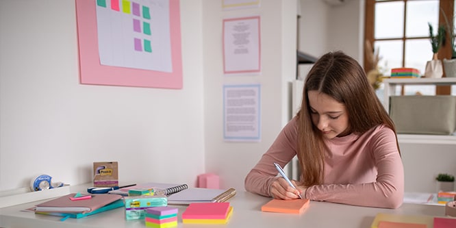 Étudiant prenant des notes sur des notes Post-it® dans un espace d'étude équipé de divers articles de papeterie, notamment des notes Post-it® et des cahiers
